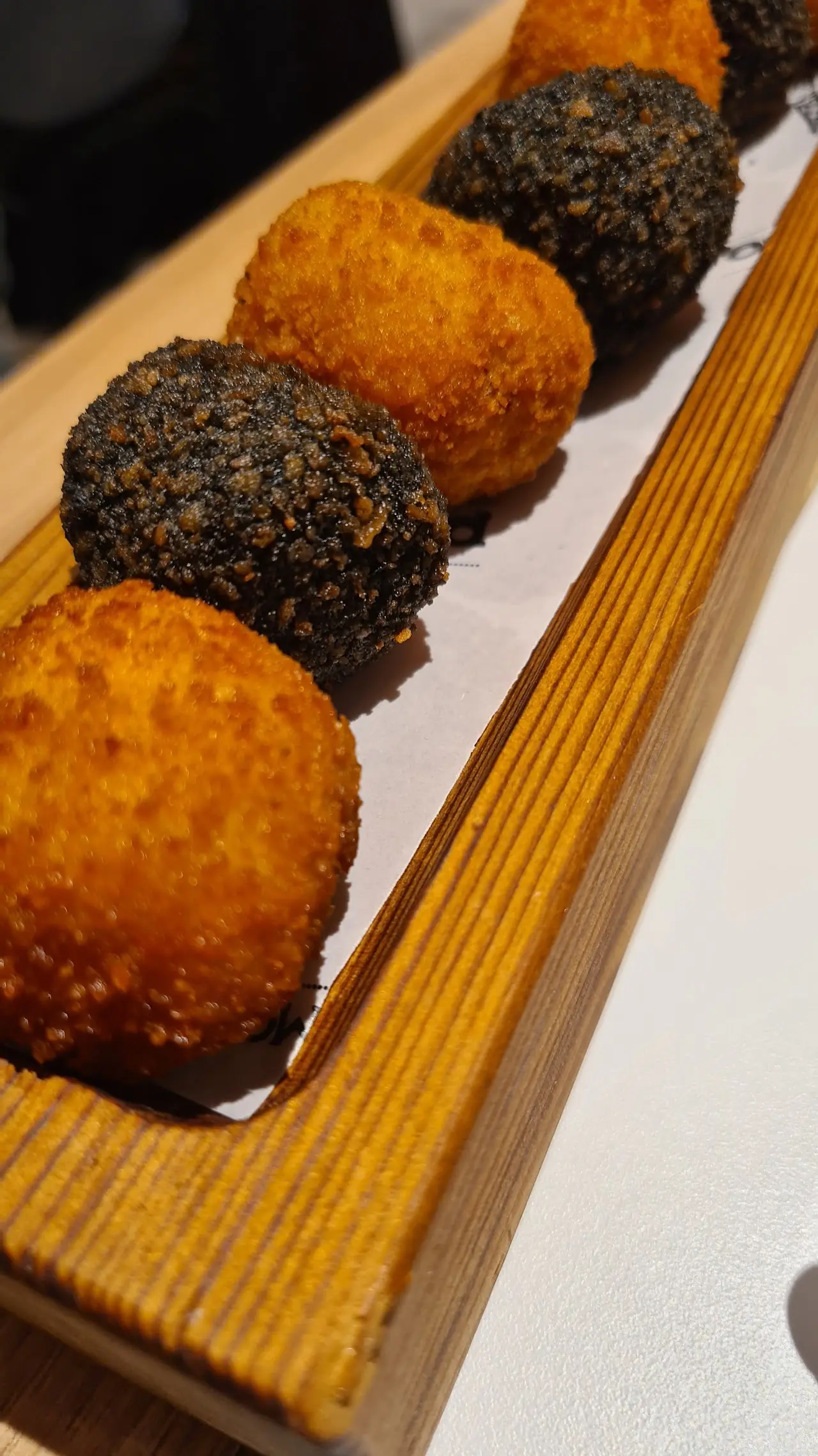 A row of fried croquettes coated in different crumbs, from golden to black, on a wooden board