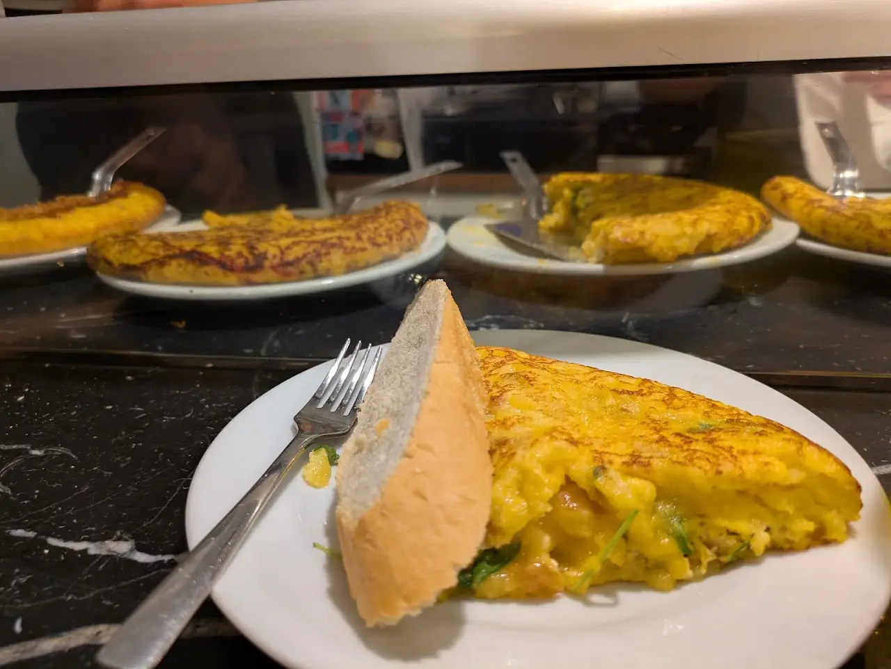 A slice of runny tortilla with bread on a white plate at the bar, more tortillas on the counter behind