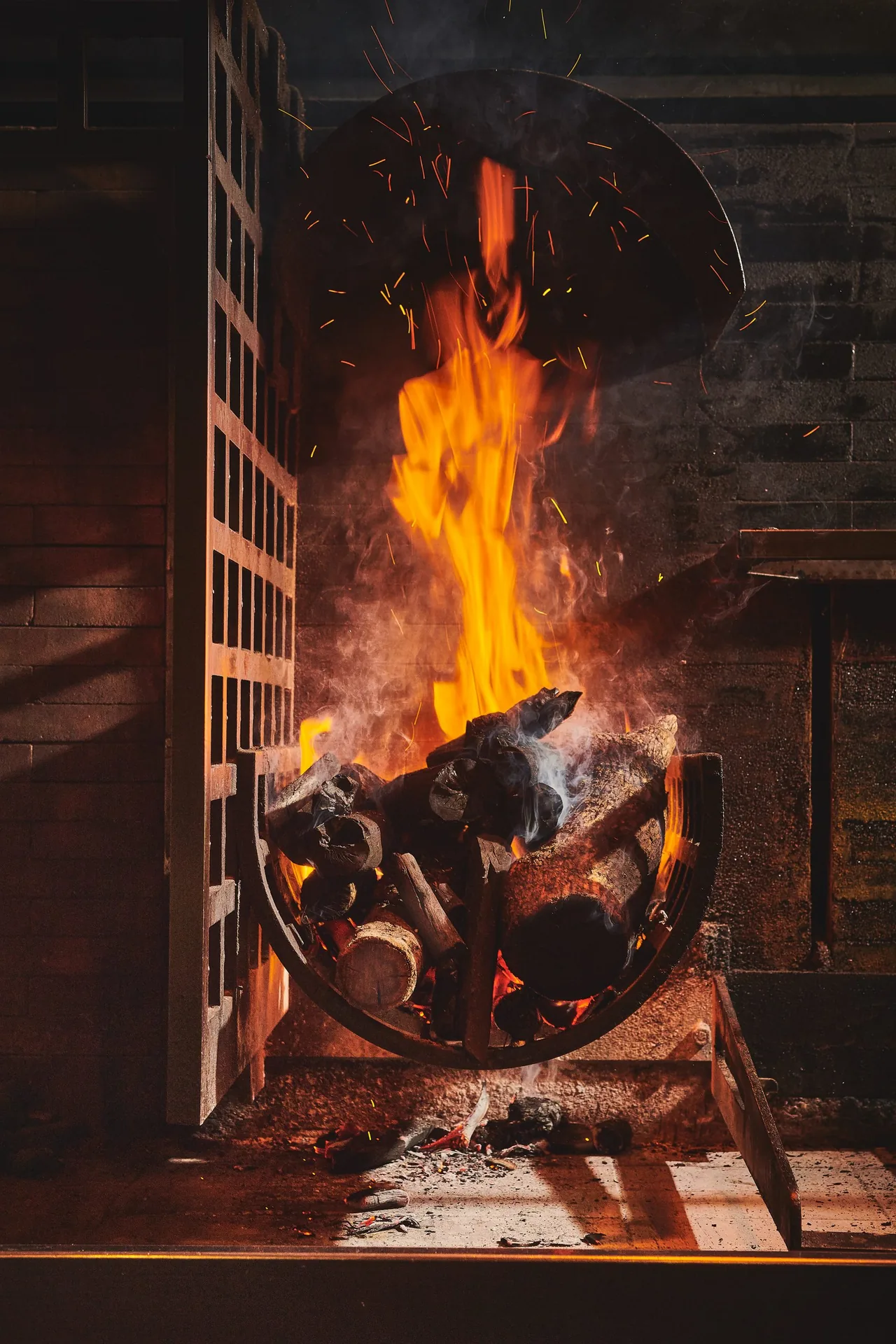 A tall wood fire burning in a semicircular iron basket inside a brick grill hearth, sparks rising past a steel hood