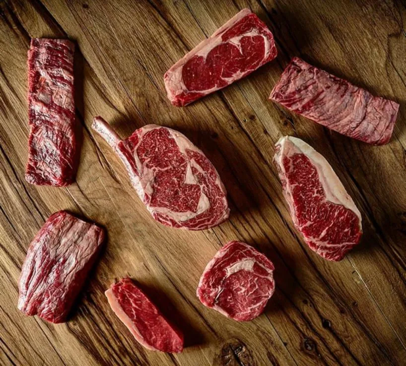 Eight raw beef cuts arranged on a weathered wooden board seen from above, including a bone-in ribeye, sirloin and skirt