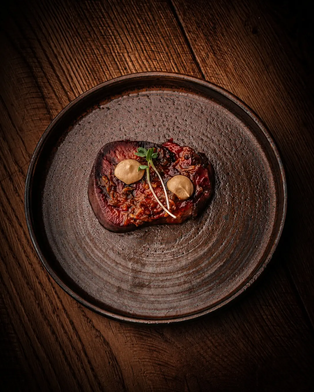 Grilled steak slice topped with a savory sauce and two dollops of creamy sauce with a microgreen garnish, on a dark ceramic plate