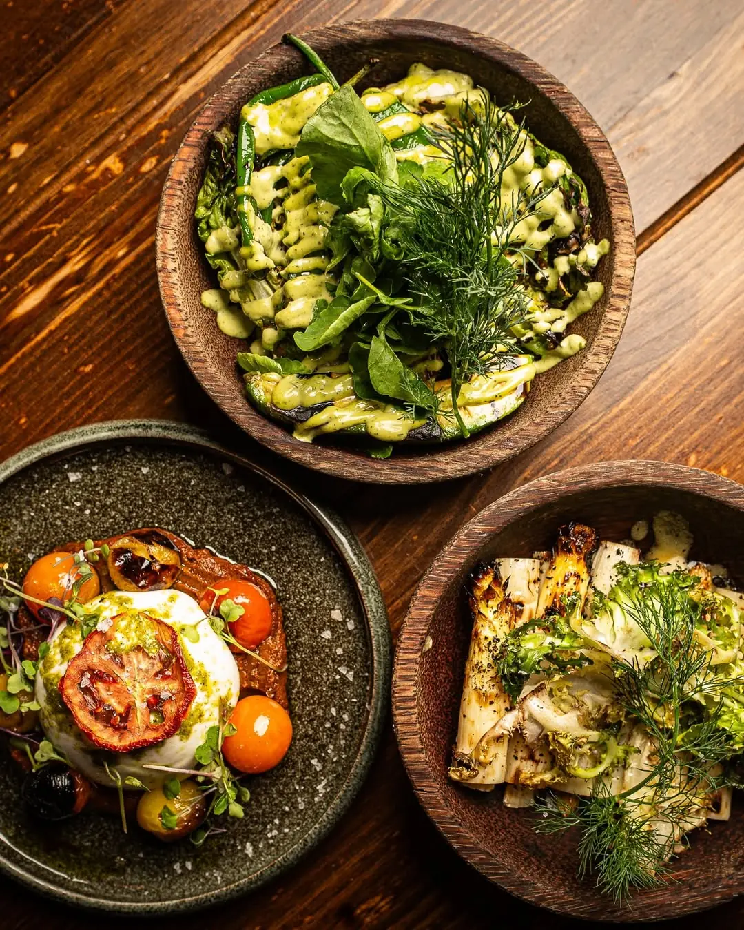 Three plated dishes on a wooden table: grilled green peppers with herb sauce, a burrata with roasted cherry tomatoes, and grilled leeks with herbs