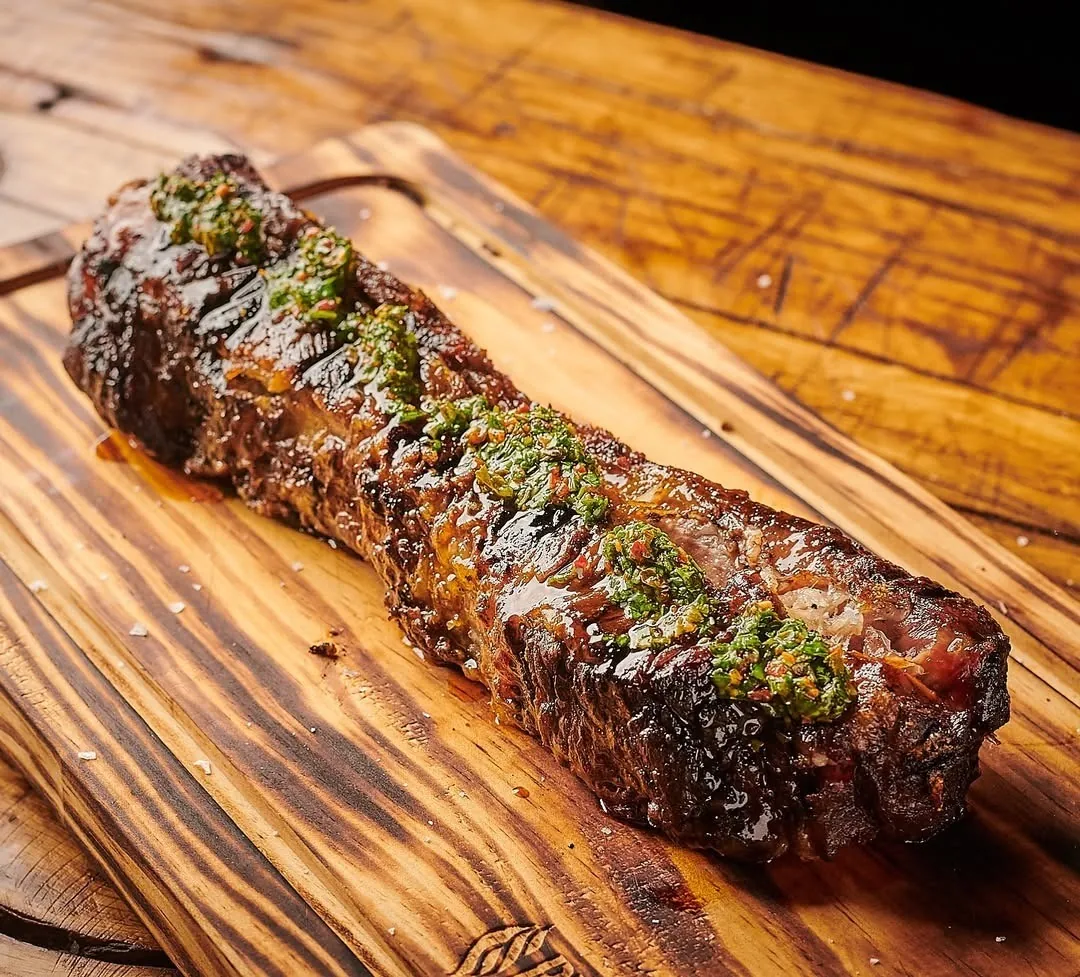 A grilled cut of beef topped with chimichurri on a charred wooden board