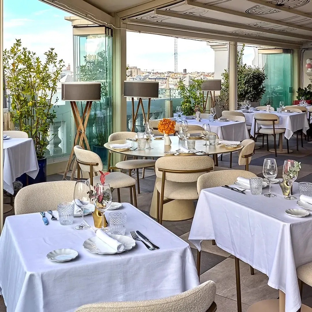 Round tables with white tablecloths and cane chairs in a glass-walled dining room with a city view