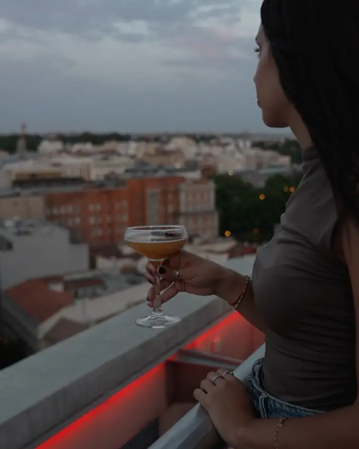 Woman holding an orange cocktail while looking out over the rooftop city view at dusk