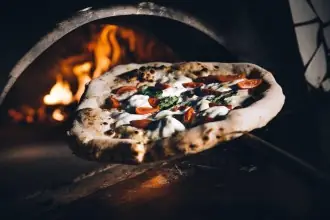 Neapolitan pizza on a metal peel being slid into a wood-fired oven with flames behind