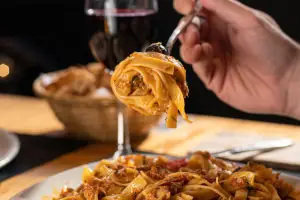Fork twirling tagliatelle in tomato ragu lifted above a plate, glass of red wine behind