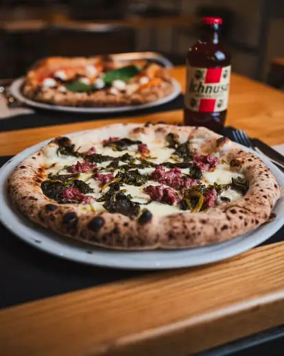 Neapolitan pizza topped with sausage and greens on a white plate, a second pizza and a bottle of Ichnusa beer behind