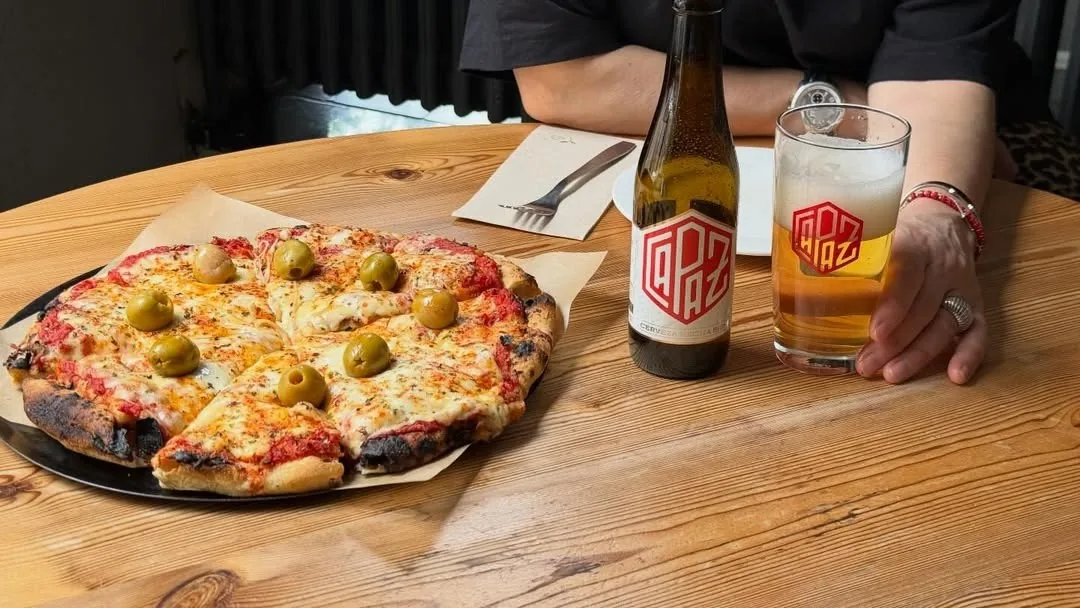 A cheese pizza topped with green olives beside a glass of beer