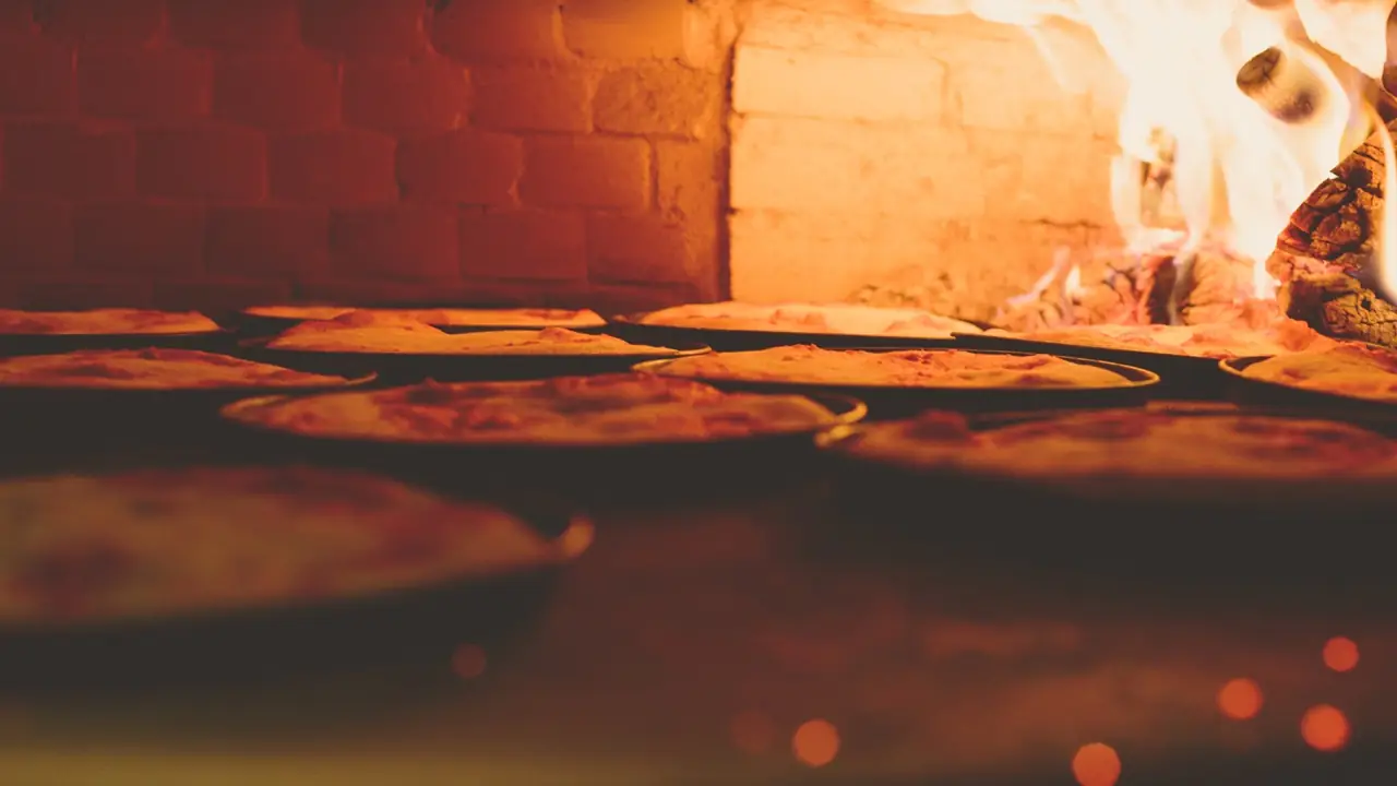 Round pizzas baking in metal pans inside a wood-fired brick oven with visible flames