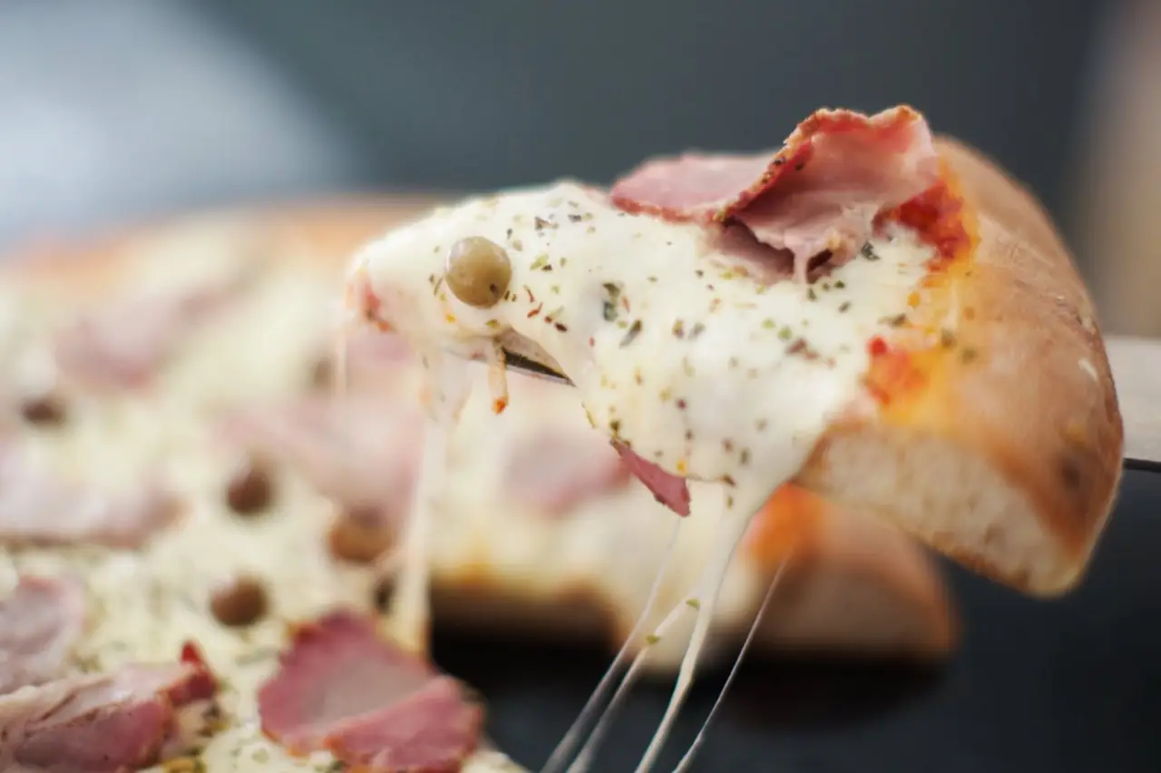Close-up of a pizza slice being lifted with stretching melted cheese, ham and capers