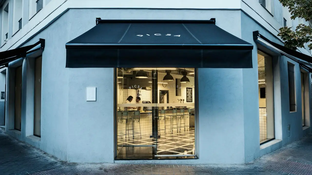 Corner storefront of PICSA with a black awning and lit interior showing a white-tiled counter and stools