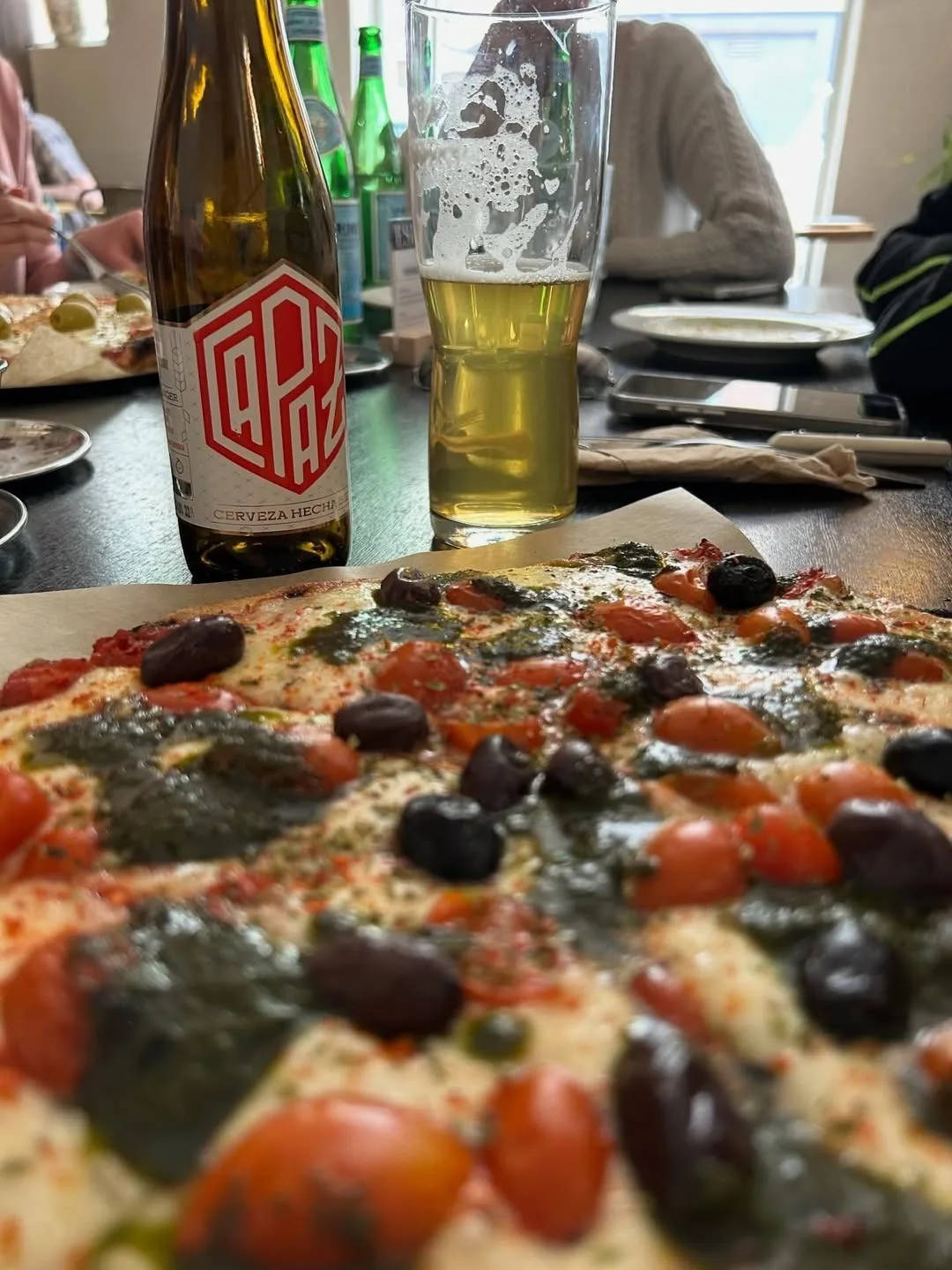A pizza topped with tomatoes, olives and pesto beside a beer