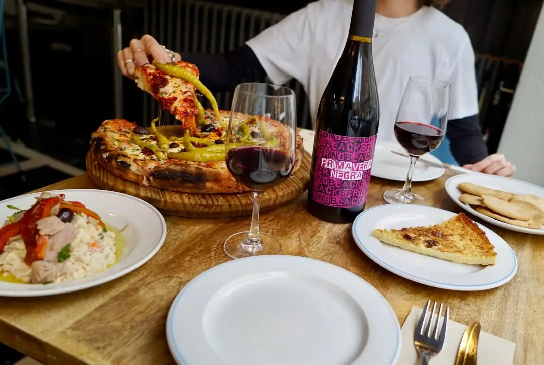 Hand pulling a cheesy slice from a whole pizza topped with green chiles and olives, on a board next to plates of stuffed peppers, bread, and glasses of red wine