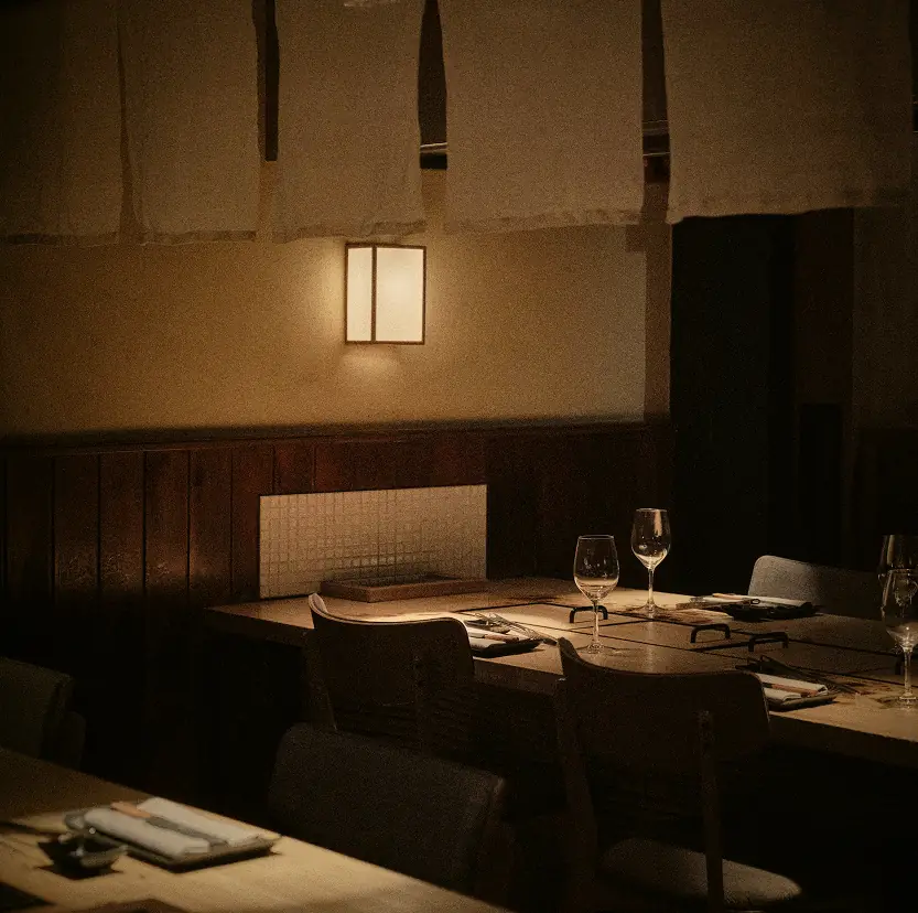 Dim Japanese dining room with linen noren panels hanging from the ceiling, a paper wall lamp on plaster above dark wood panelling, and wooden tables laid with wine glasses, chopsticks and small dishes