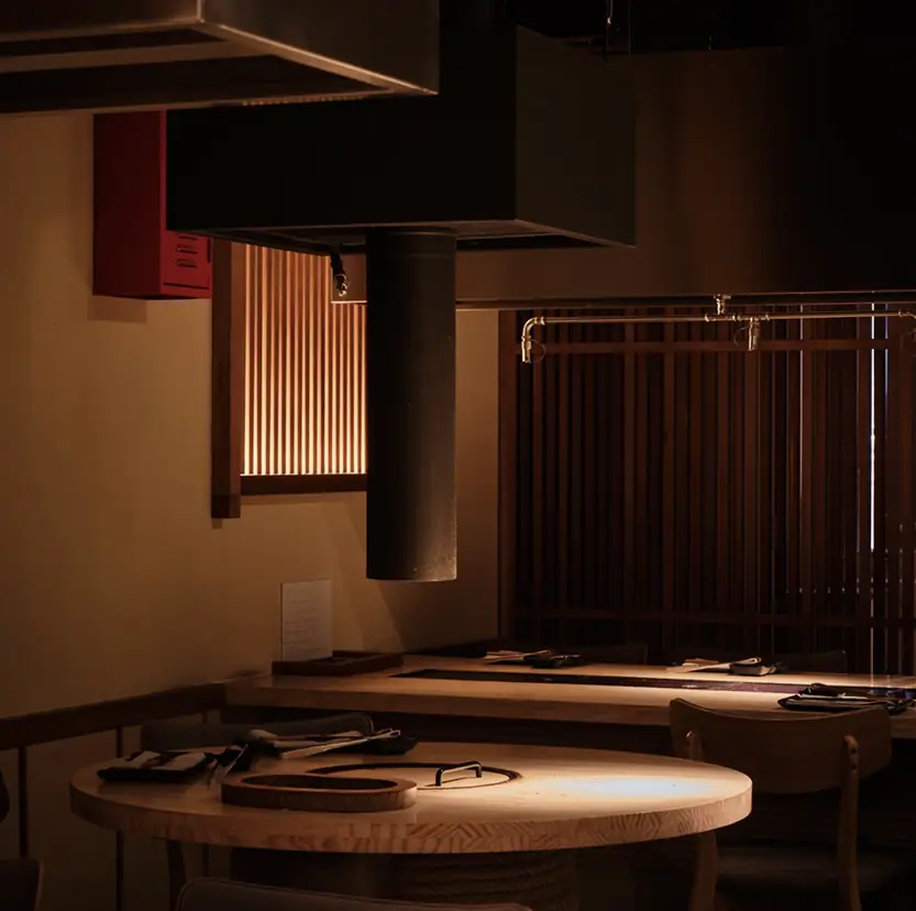 Low-lit grill room: a square extraction hood on a metal flue hangs over a wooden counter with a recessed charcoal grill, backed by slatted wood screens and a warm slatted light panel
