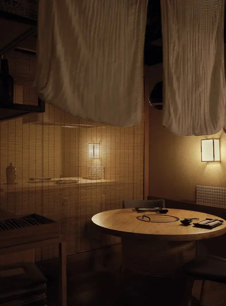 Corner of the restaurant lit by a small paper wall lamp, with a round pale wood table holding a recessed grill plate, small black dishes and chopstick rests, framed by hanging cream cloth panels and a bamboo screen