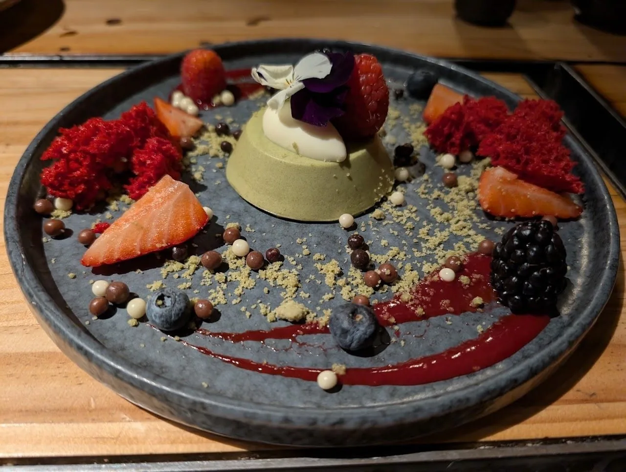 A green dome dessert on a dark plate surrounded by berries and a sauce swirl