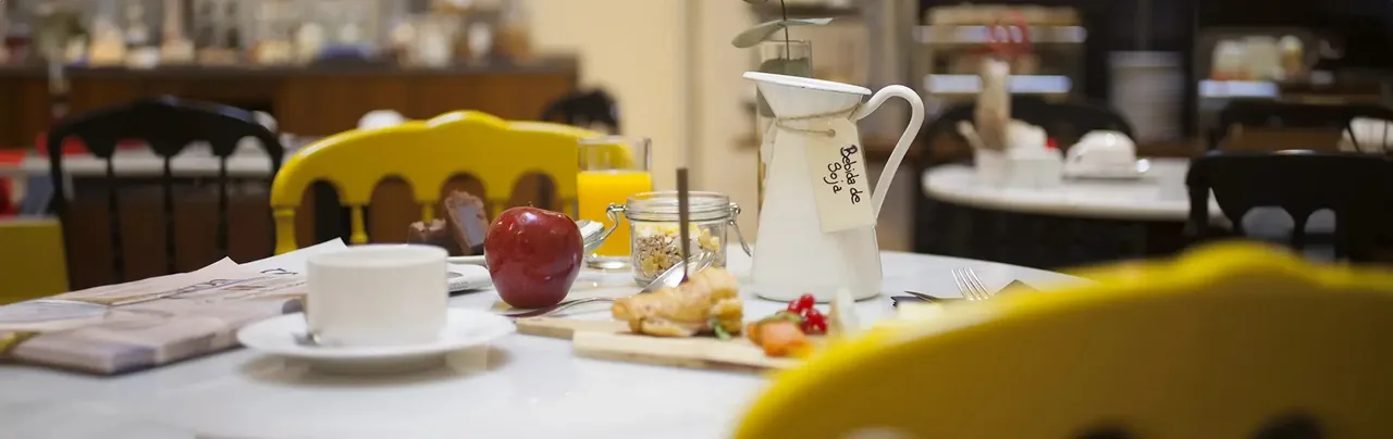 Breakfast laid on a marble table: a coffee cup and saucer, a red apple, orange juice, a kilner jar of granola, pastries on a wooden board and an enamel jug tagged Bebida de Soja, with yellow spindle-back chairs around