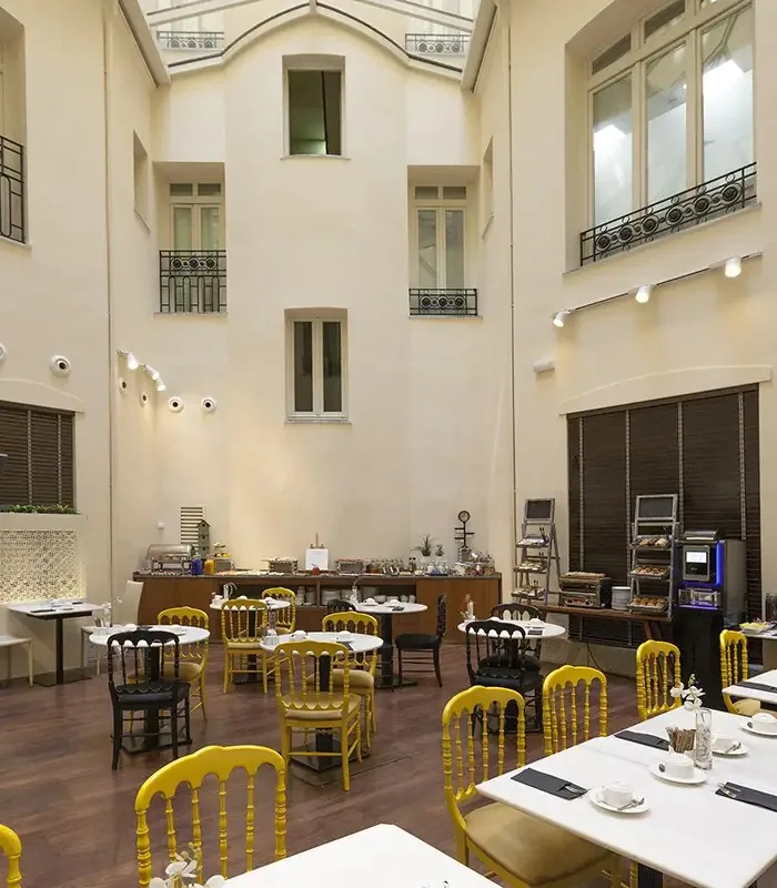 Tall glass-roofed atrium breakfast room with cream walls and wrought-iron balconies overhead, yellow spindle-back chairs at round marble tables and a buffet counter with chafing dishes and coffee machines along the back