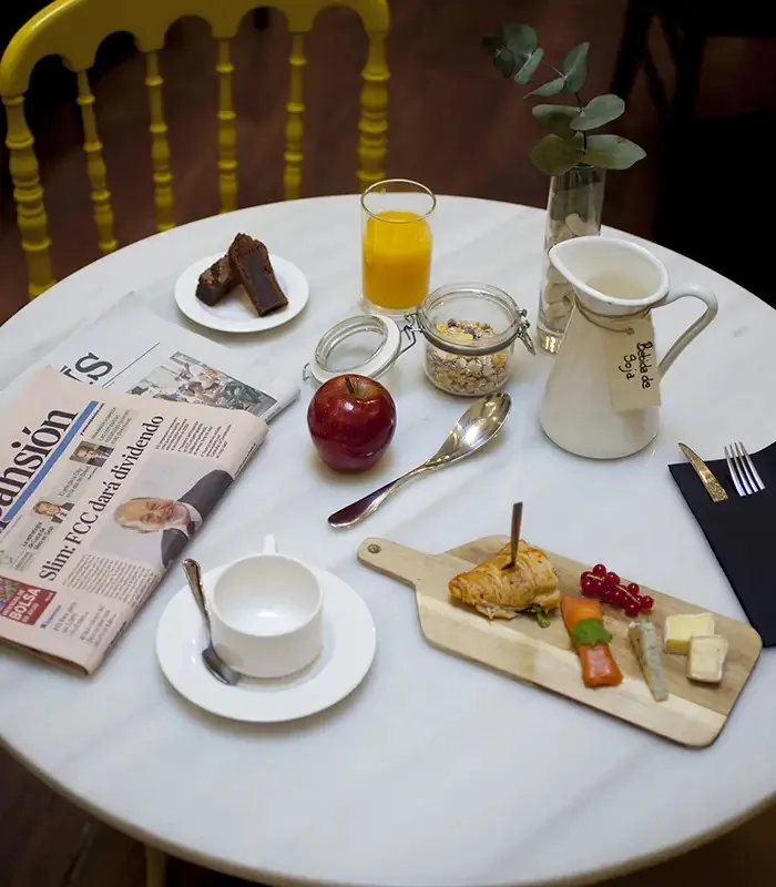 Overhead of a breakfast table with a filled croissant, cheese, redcurrants and carrot on a wooden board, brownie squares, orange juice, granola jar, apple, soy-milk jug and a folded Expansion newspaper