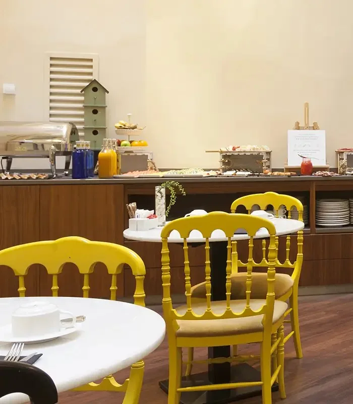 Breakfast buffet counter with chafing dishes, bottled juices, cured meats, pastries and a tiered fruit stand, yellow chairs at marble tables in front and stacked plates below
