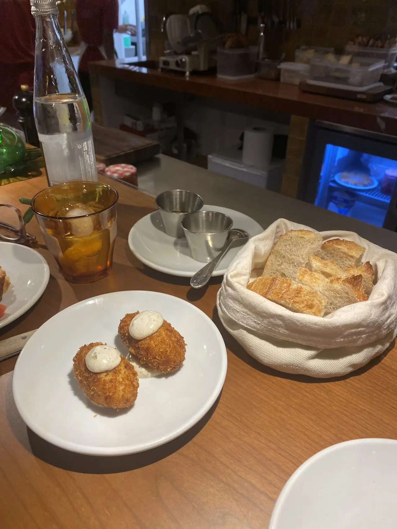 Two croquettes topped with aioli on a white plate beside a basket of bread, at Piropo Bistro.