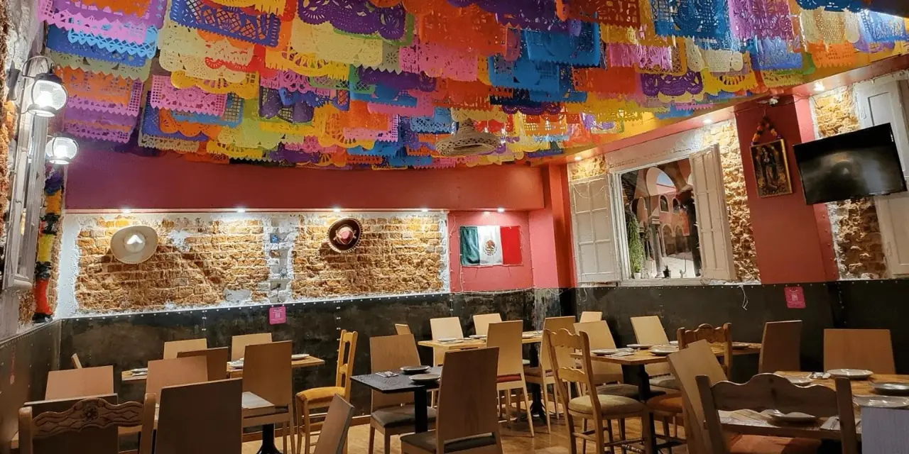 Mexican restaurant dining room with colorful papel picado banners across the ceiling, exposed brick walls, a Mexican flag, wooden tables and chairs