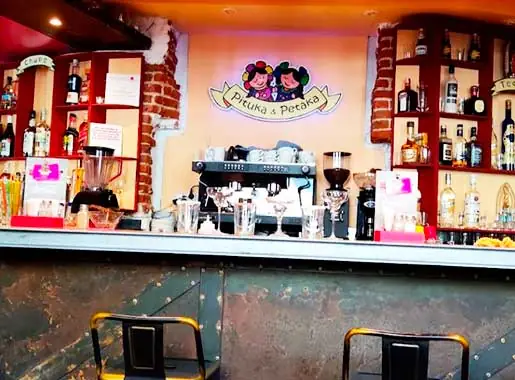 Bar counter with espresso machine, blenders and rows of bottles beneath a wall-mounted 'Pituka & Petaka' logo, with two stools in front