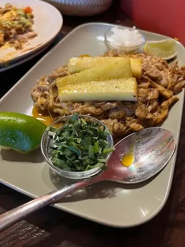 Plate of shredded meat topped with cheese-filled rolled tortillas, with lime wedges, a dish of chopped cilantro and a spoon