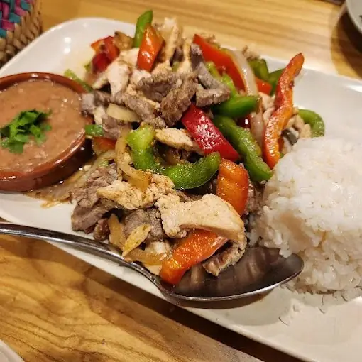 White plate of stir-fried beef and chicken with red and green peppers and onions, served with white rice and a bowl of refried beans