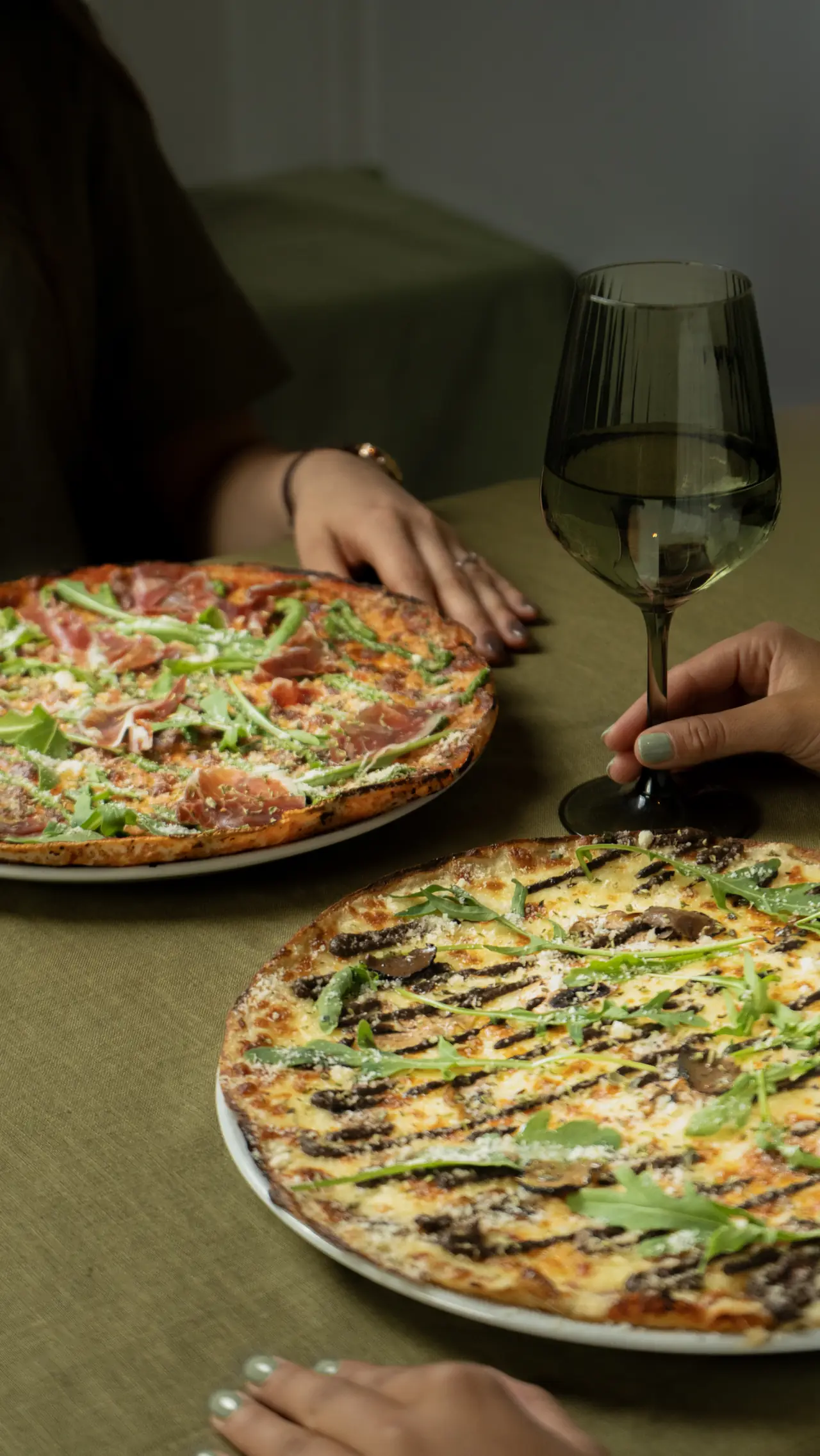 Two grilled-crust pizzas on white plates on a table, one topped with prosciutto and arugula, the other with mushrooms and arugula, at Pizza Natura