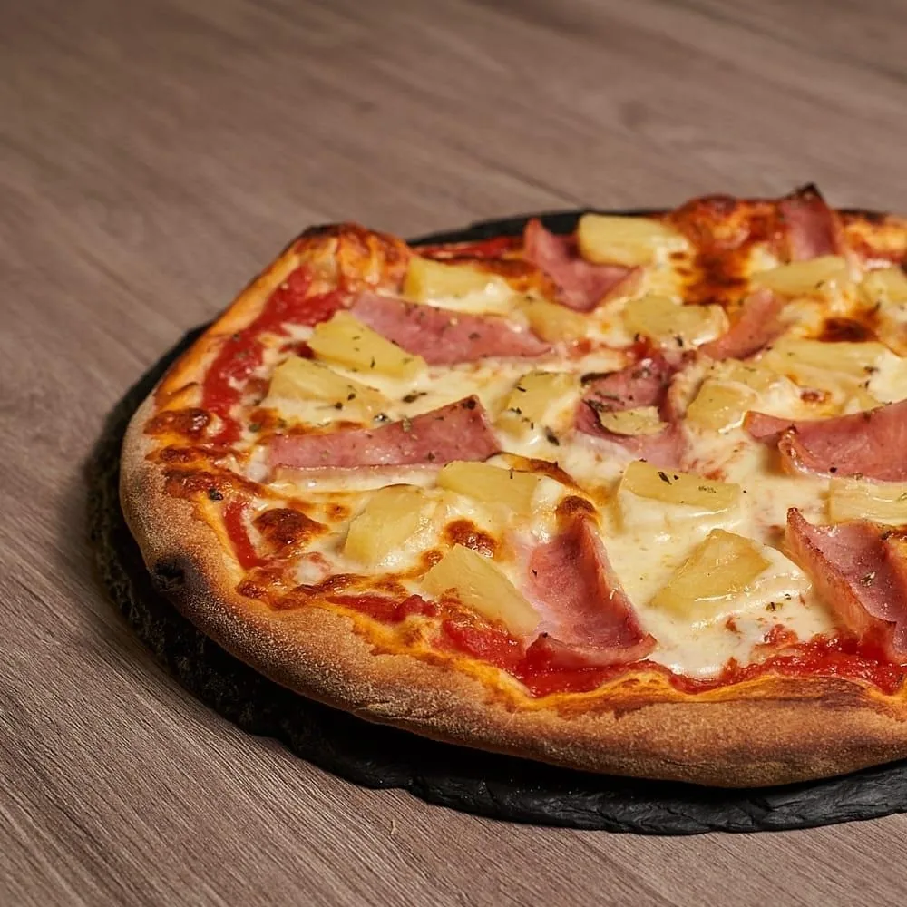 A pizza with ham and pineapple on a dark slate board on a wooden table