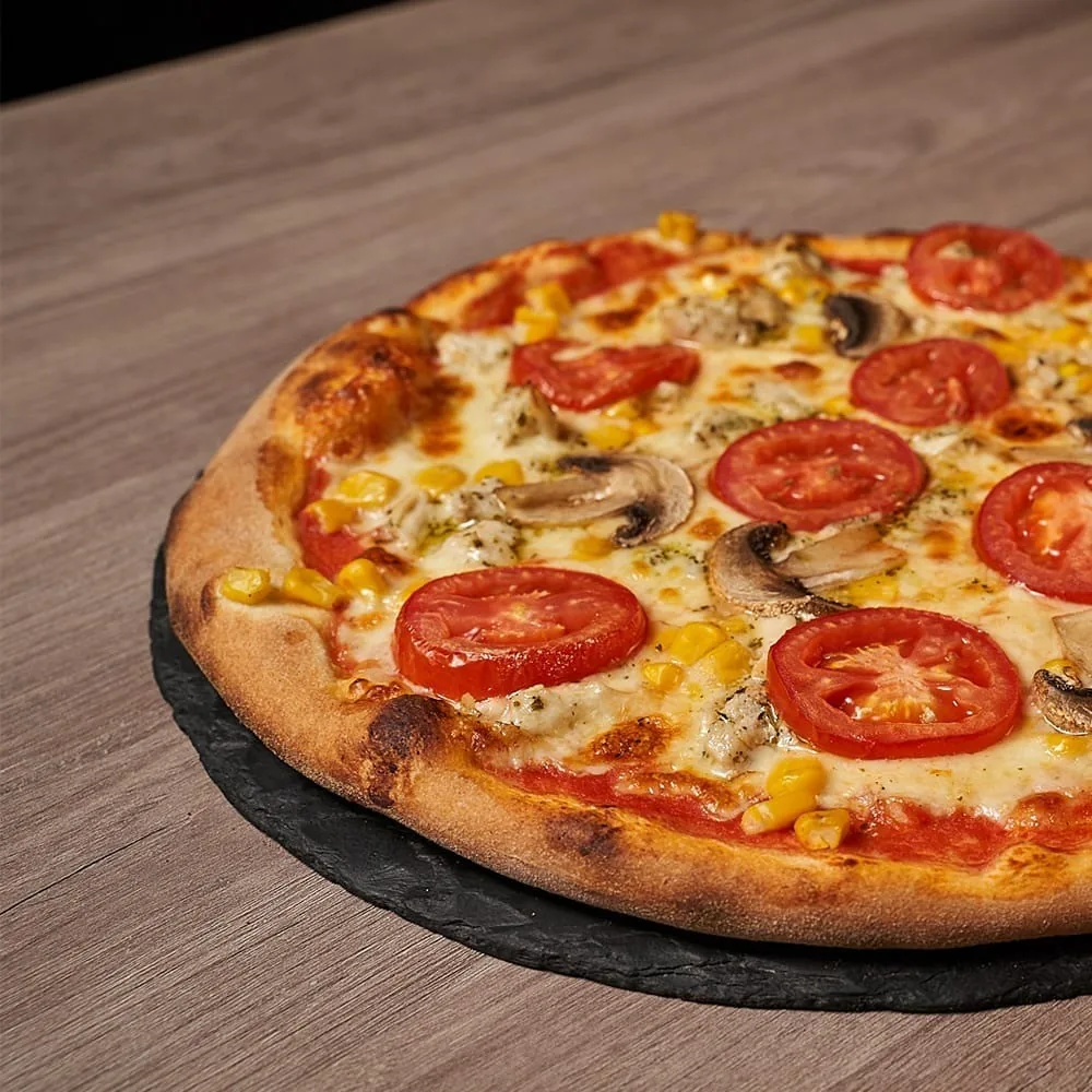 A pizza with tomato slices and sweetcorn on a dark slate board on a wooden table