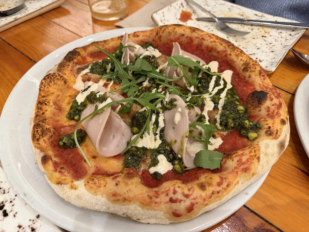 A pizza with ham slices, rocket and a white drizzle on a white plate on a wooden table