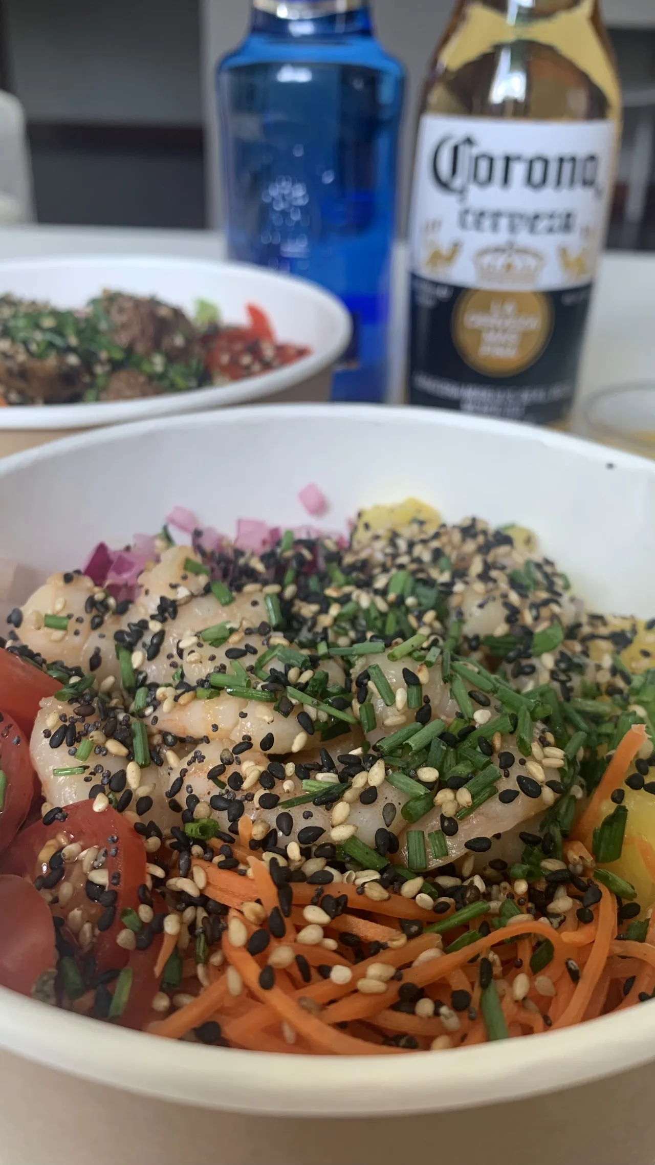 A poke bowl topped with greens, carrots, and sesame seeds, beer bottle beside it