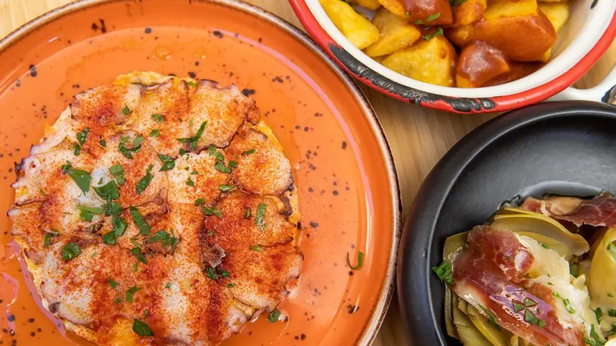 Octopus with paprika on orange plate, alongside potatoes and artichoke with jamon