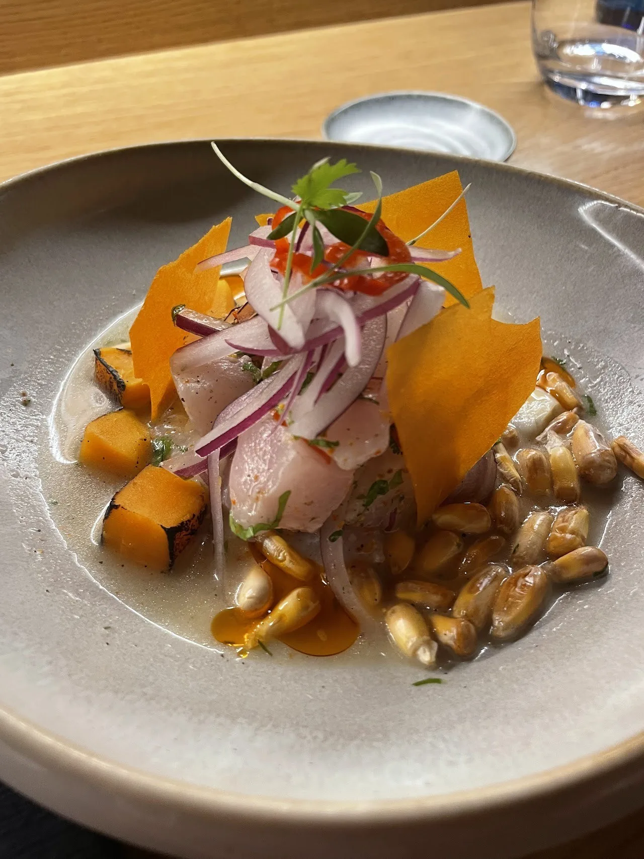 A grey bowl of ceviche with red onion, orange sweet potato, toasted corn and a crisp garnish
