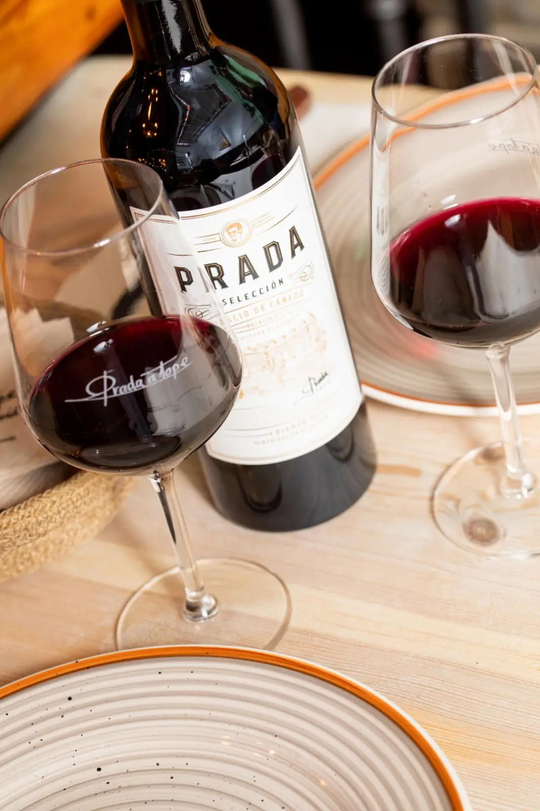 Bottle of Prada Seleccion Palacio de Canedo red wine flanked by two poured glasses of red wine on branded 'Prada a Tope' stemware and rimmed plates