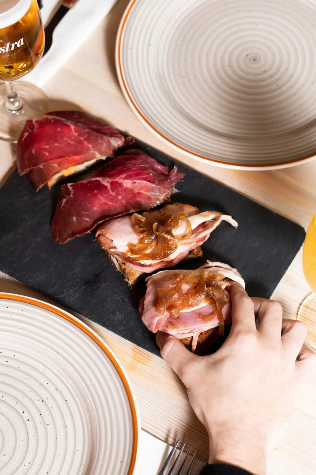 Slate board of thin-sliced cured beef cecina and open toasts topped with pork loin and caramelised onion, a hand reaching for one, with a beer glass alongside