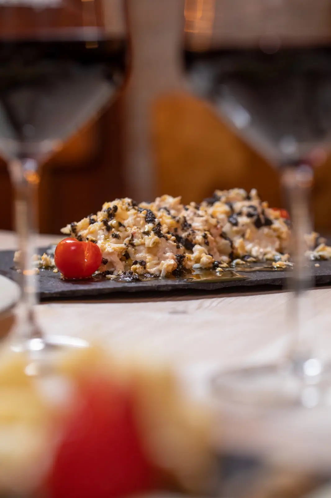 Plated scrambled-egg revuelto topped with dark morcilla or truffle on a slate, garnished with a cherry tomato, framed by out-of-focus wine glasses