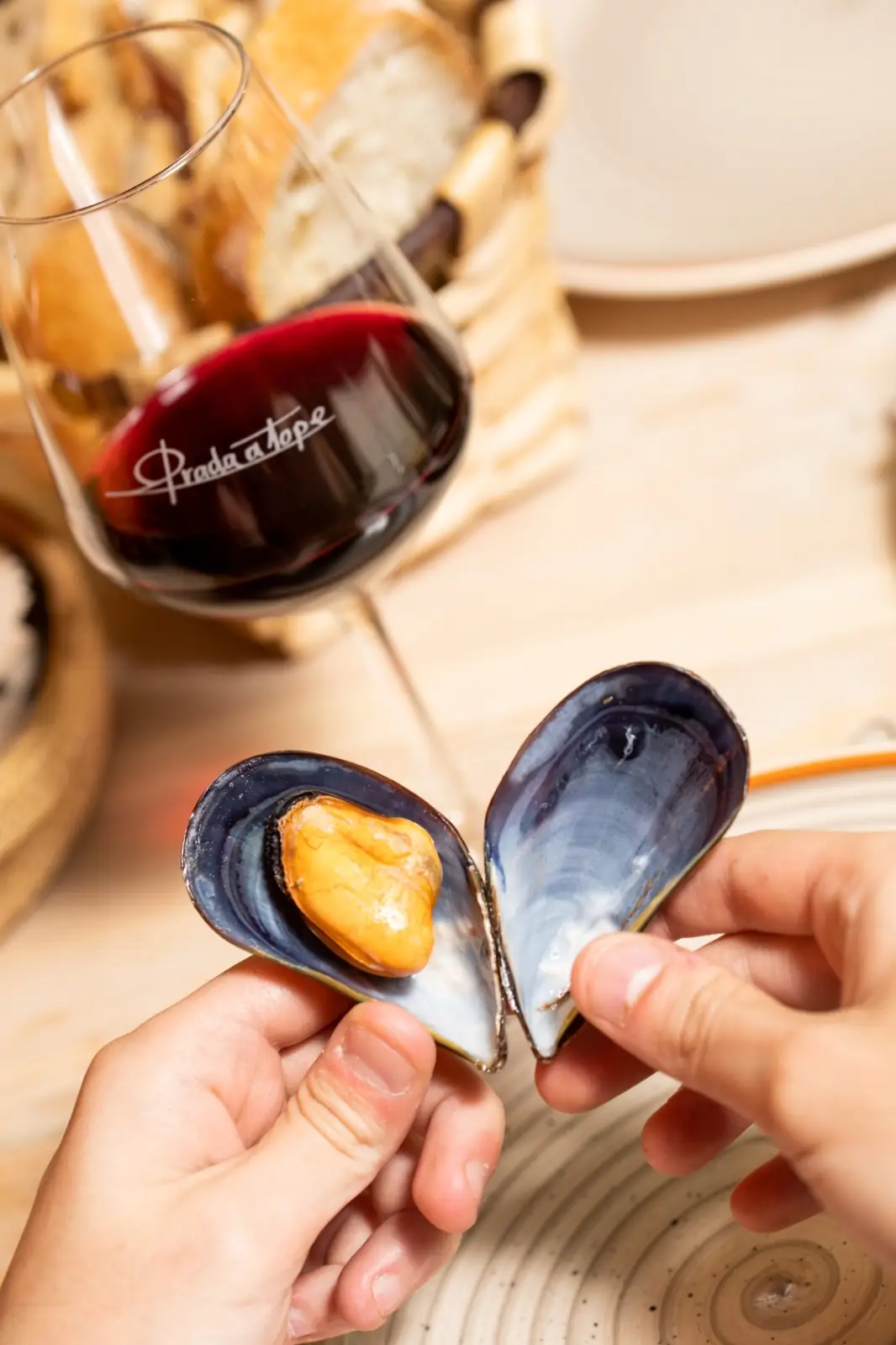 Two hands holding open a cooked mussel shell to reveal the orange meat, with a branded 'Prada a Tope' glass of red wine and a bread basket behind