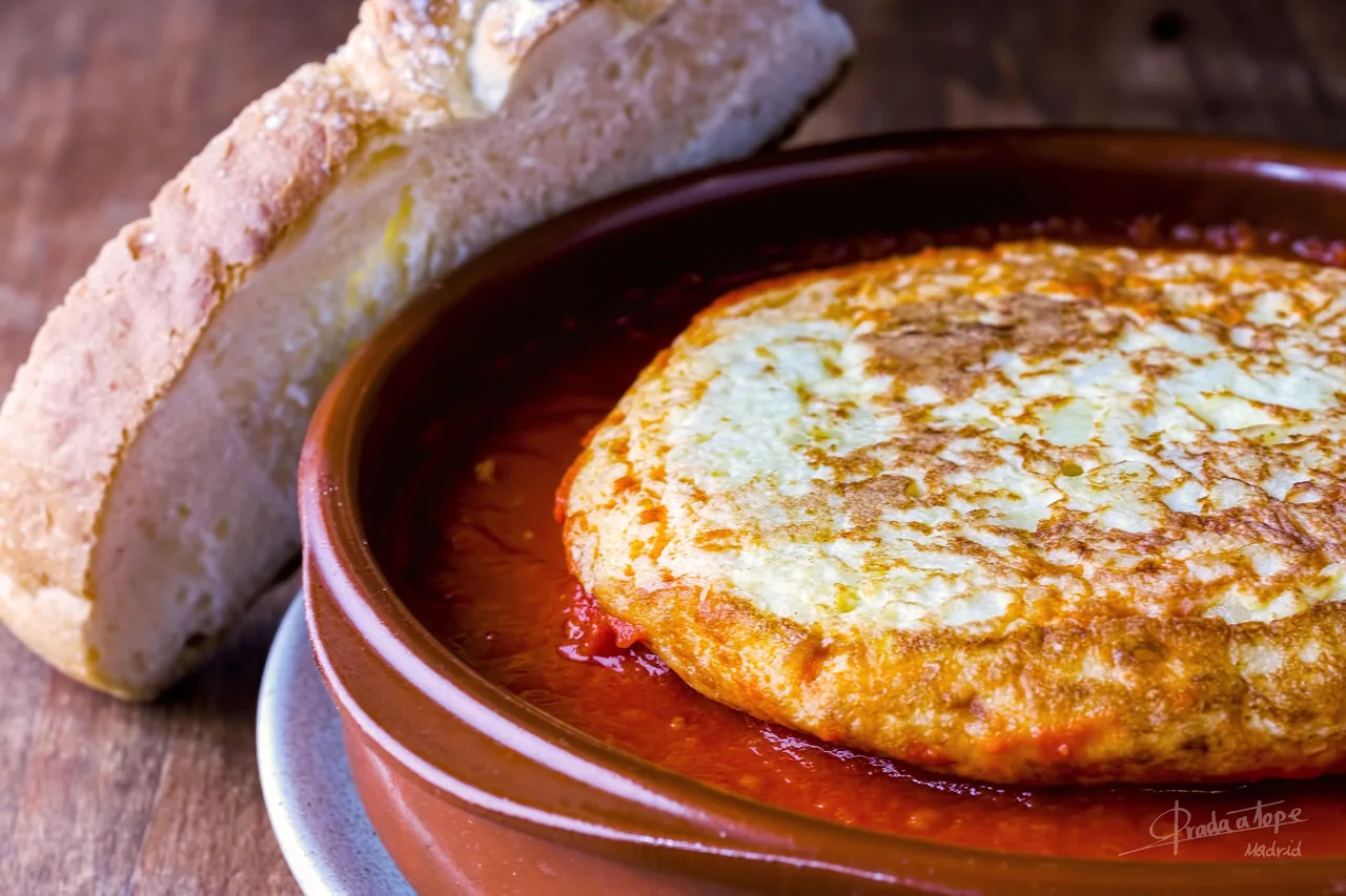 A thick golden omelette in a brown clay dish beside a piece of bread at Prada a Tope