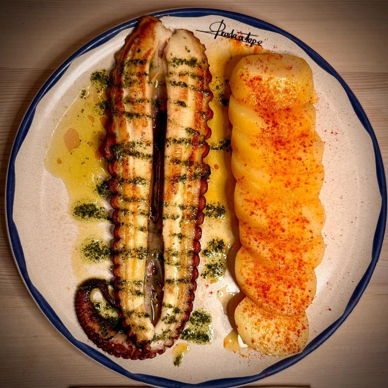 A white plate with a grilled octopus tentacle and a golden roasted potato with green sauce
