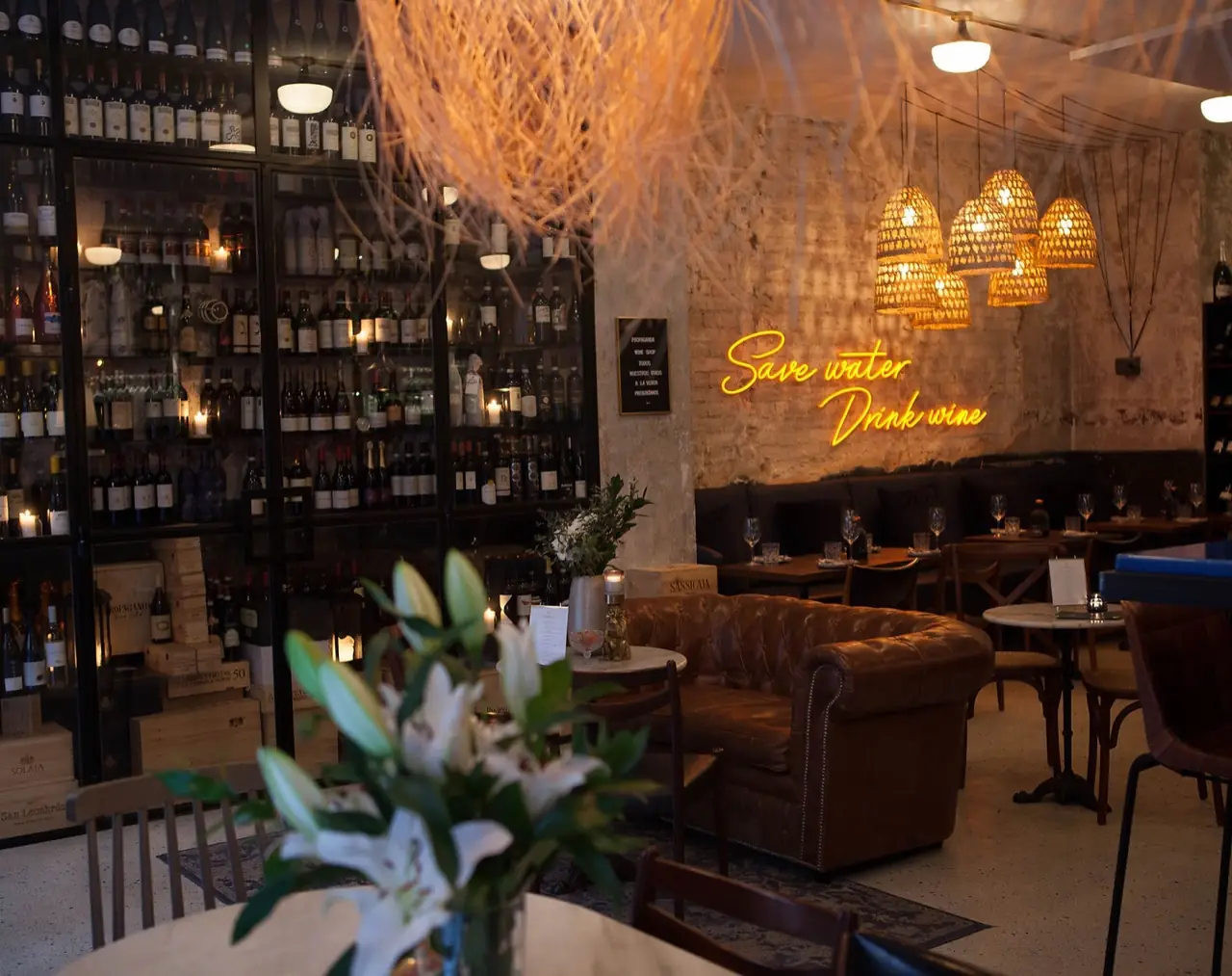 Wine bar interior with a floor-to-ceiling backlit bottle wall, a brown leather Chesterfield sofa and a yellow neon sign reading Save water Drink wine