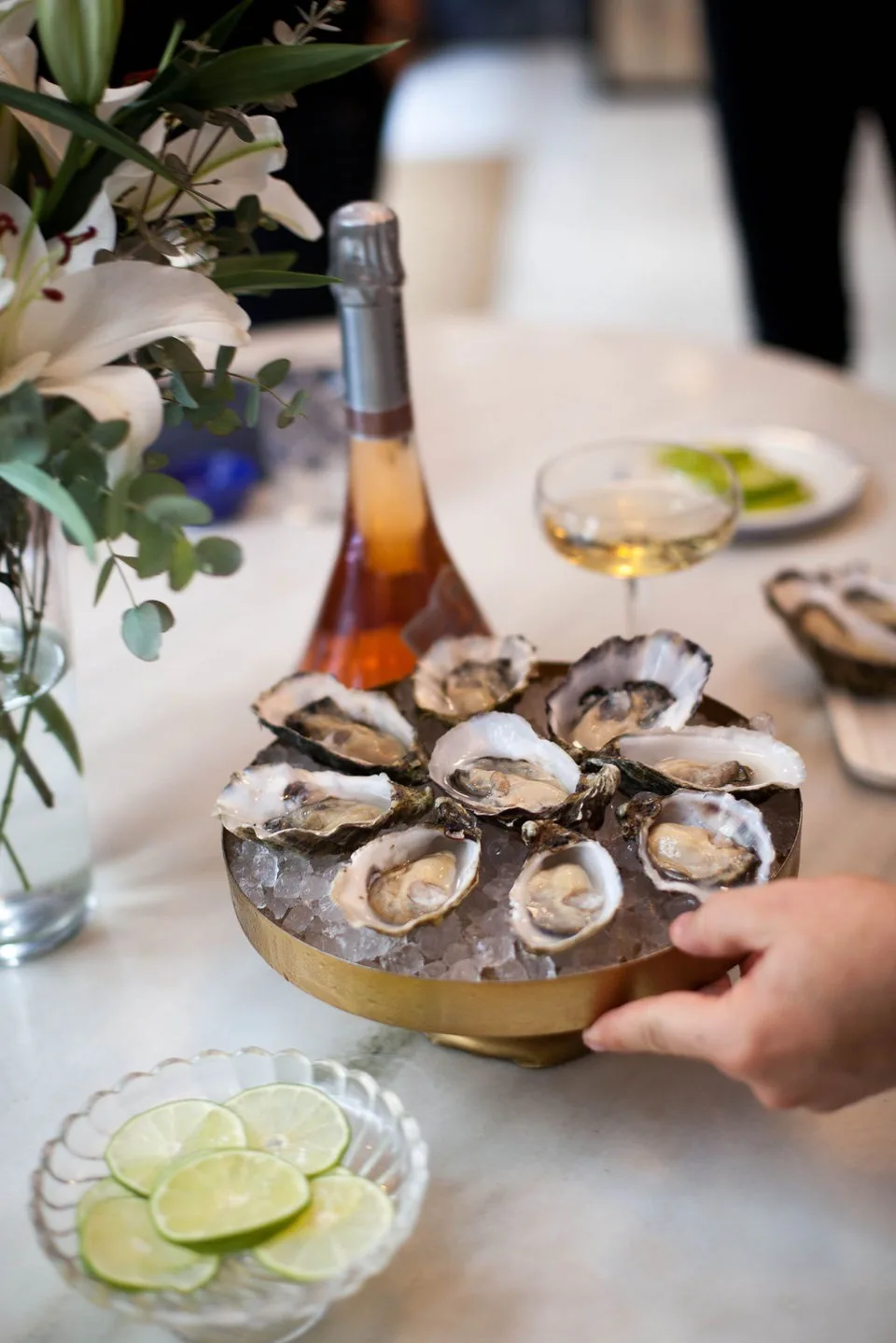 A silver tray of oysters on ice with lime wedges, held by one hand