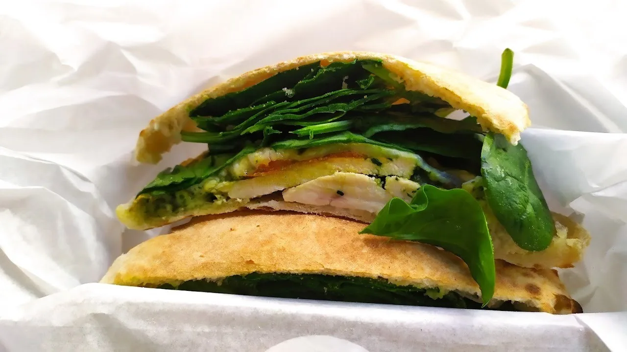 A halved flatbread sandwich stuffed with spinach leaves, sliced chicken and green sauce on white paper