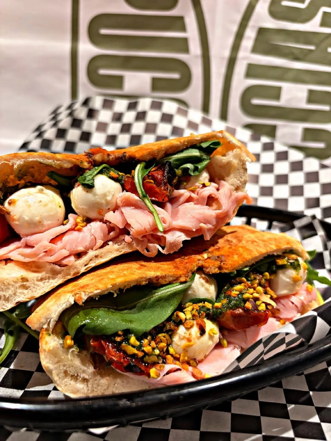 Two halves of a flatbread sandwich filled with ham, mozzarella balls, rocket, tomato and chopped pistachios in a black basket