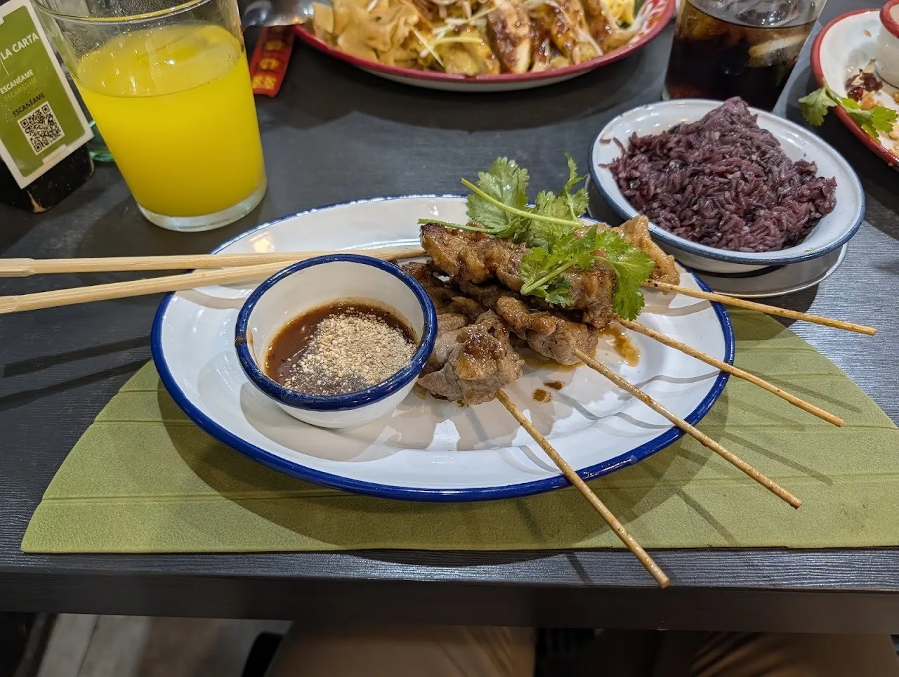Grilled meat skewers with a dipping sauce on a plate beside a bowl of dark rice