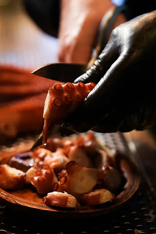 Close-up of a hand slicing a grilled octopus tentacle onto a wooden plate of sliced pulpo a feira at Pulperia Can Lampazas
